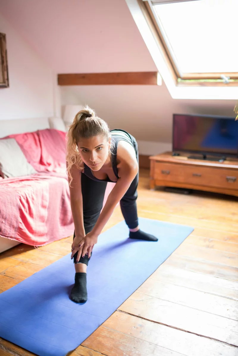 A woman performs a stretching exercise on a yoga mat in a bright, cozy room.