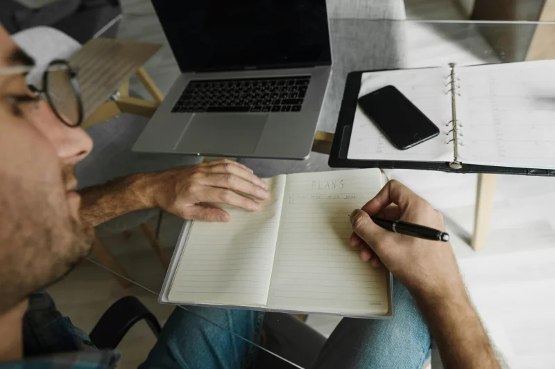 A man writes in a notebook on a glass desk, working alongside a laptop and planner.