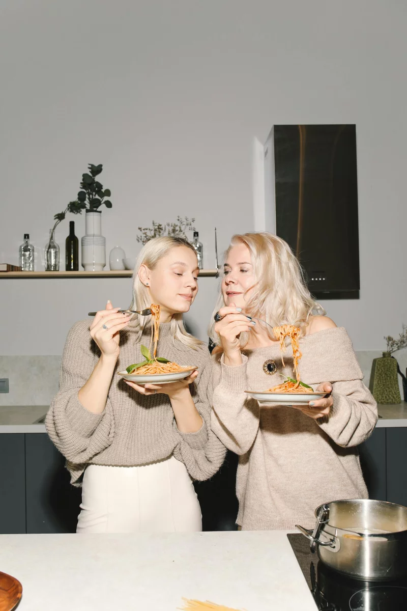 A joyful blonde mother and daughter enjoying pasta in the kitchen, showcasing family bonding and Italian cuisine.