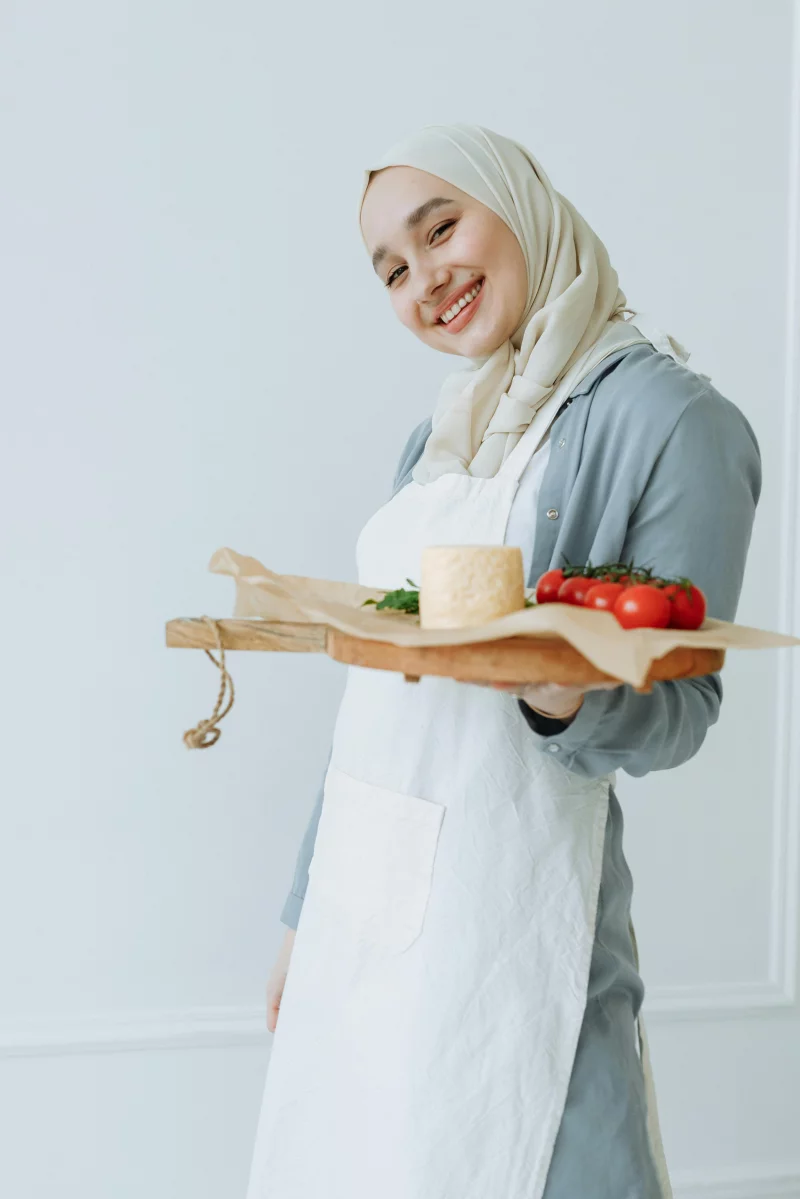 Cheerful woman wearing a hijab holding a tray with fresh tomatoes and cheese indoors.