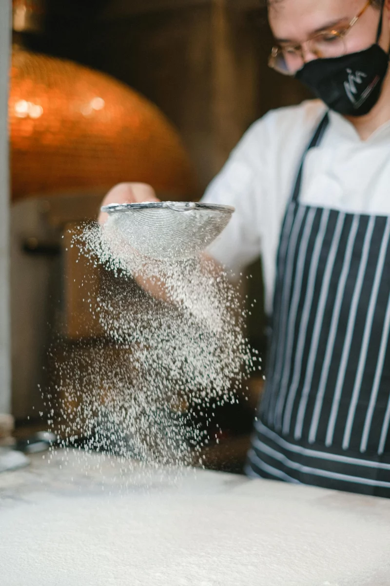 A chef wearing an apron sifts flour onto a preparation surface in a professional kitchen.
