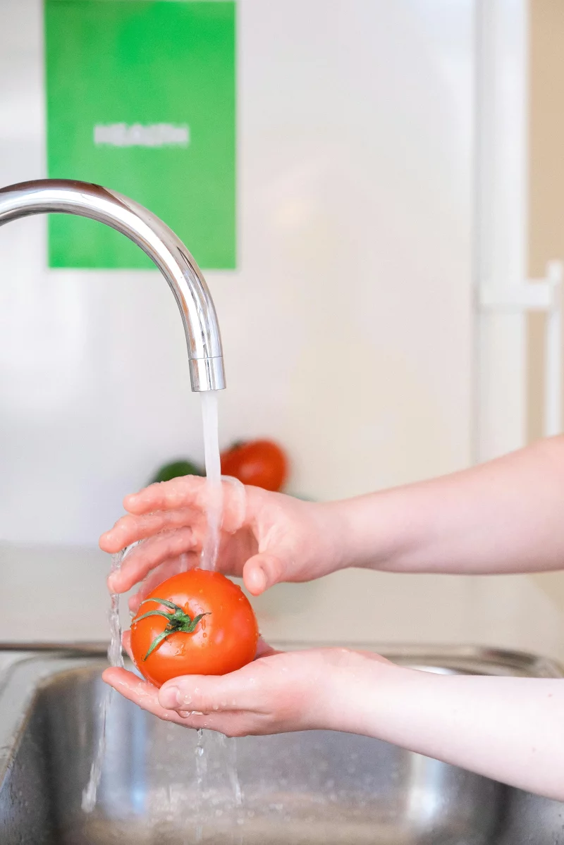 Close-up of hands washing a tomato under a kitchen faucet, promoting cleanliness and health.