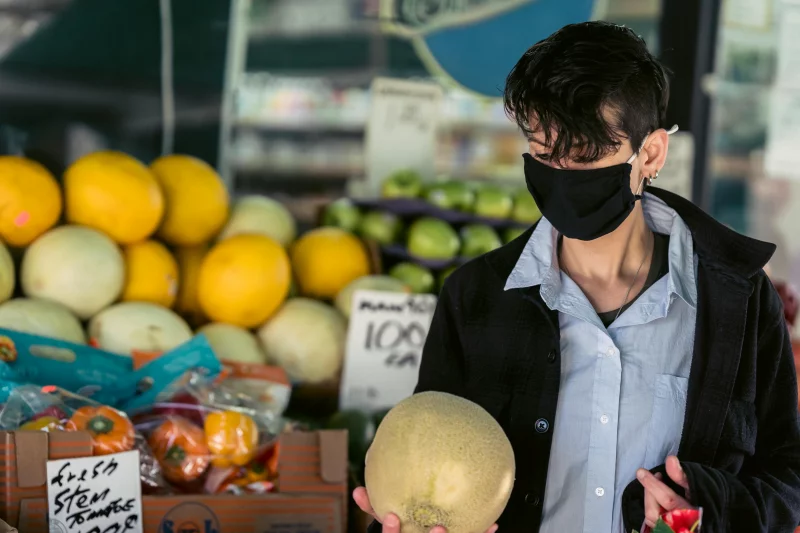 Masked individual selecting fresh melon at a vibrant outdoor market showcasing colorful fruit displays.