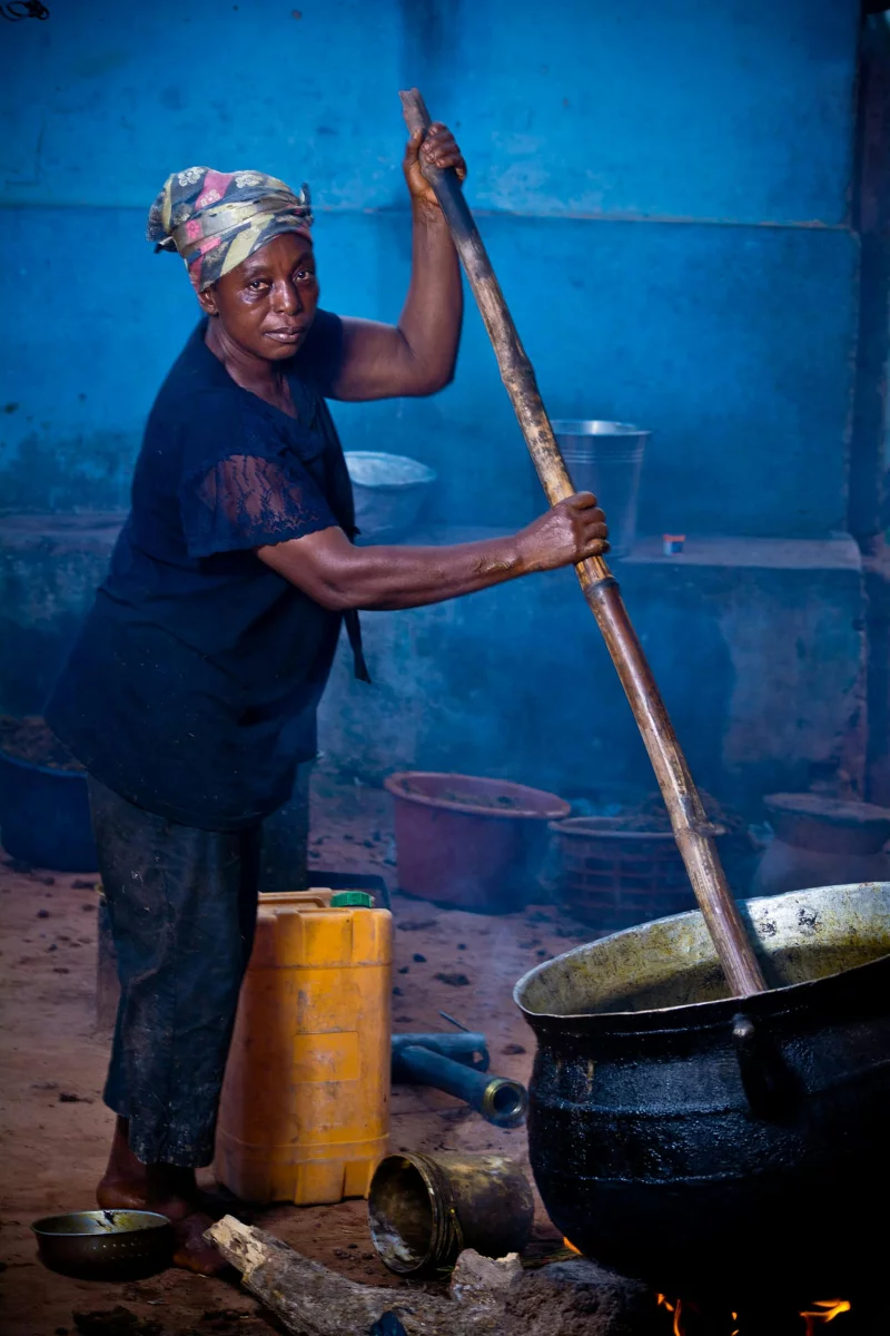 Elderly woman stirring a large pot outdoors in Ghana, showcasing traditional cooking.