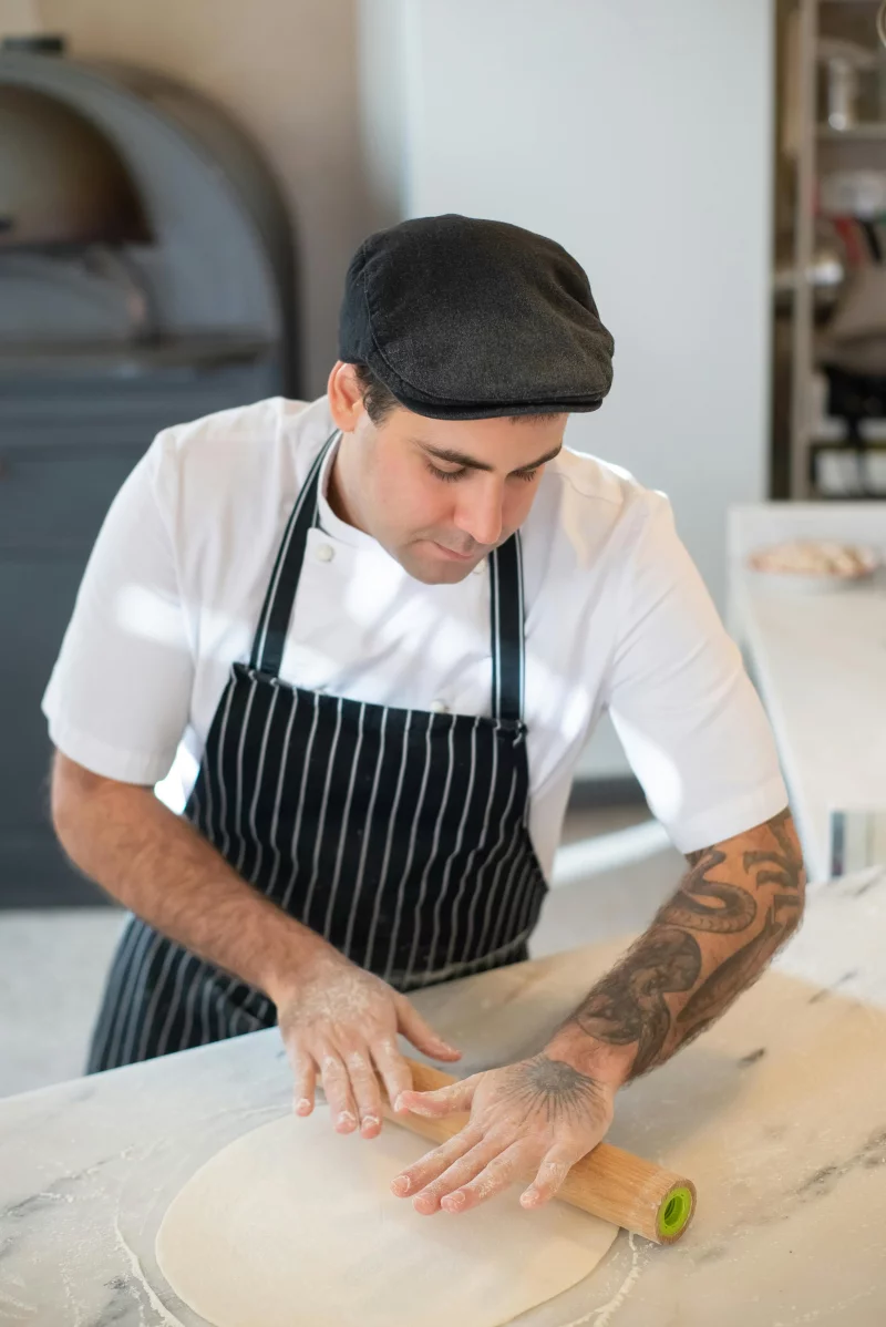 Chef expertly rolls dough in a well-lit kitchen, showcasing culinary skills and tattoos.