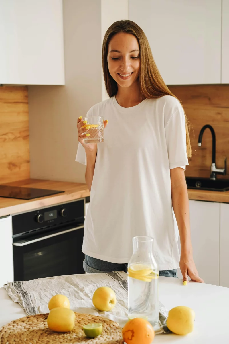 Smiling woman holding glass of lemon water in a stylish kitchen, promoting a healthy lifestyle.