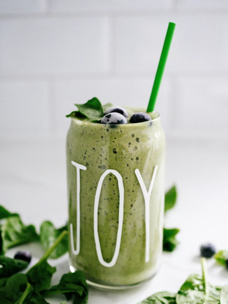 Close-up of a green smoothie with blueberries in a 'JOY' mug, garnished with fresh leaves and straw.