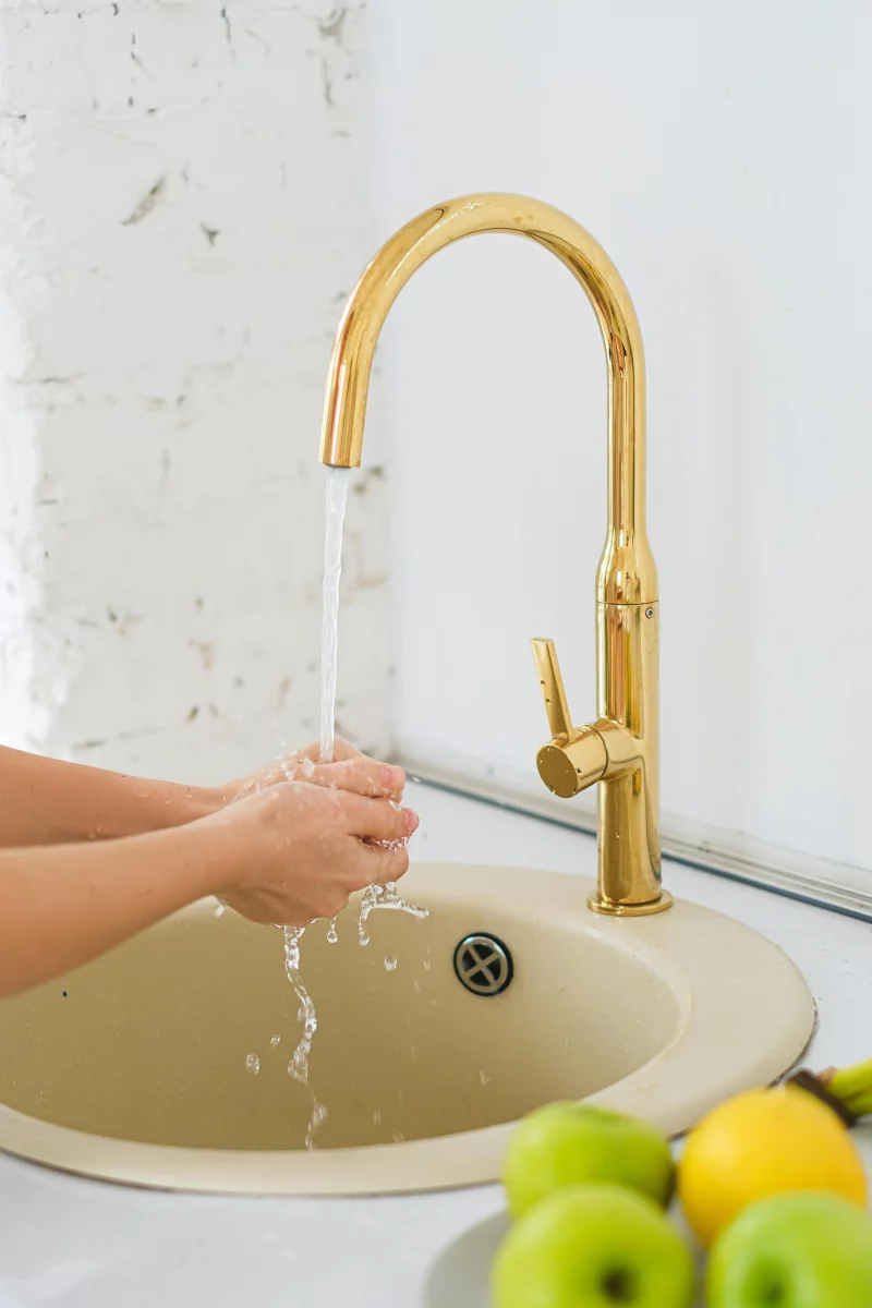 Hands washing under a stylish gold faucet with apples and lemons nearby in an indoor setting.