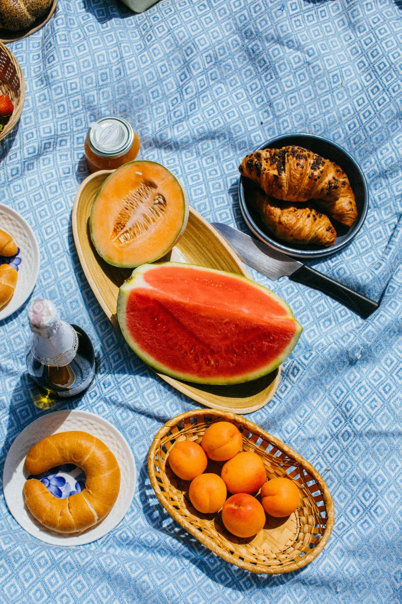 Vibrant picnic setup with melon, croissant, and apricots on blanket.