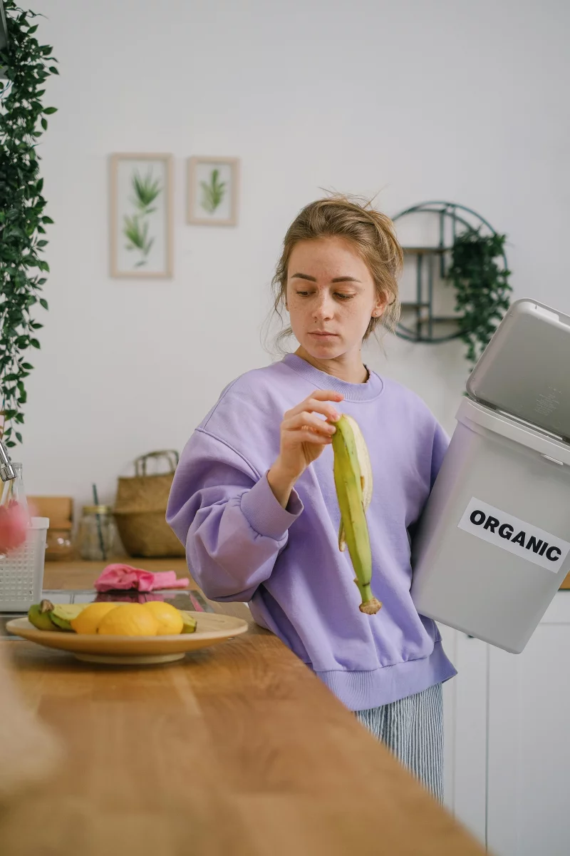 Woman disposing banana peel into an organic waste bin in a cozy kitchen.