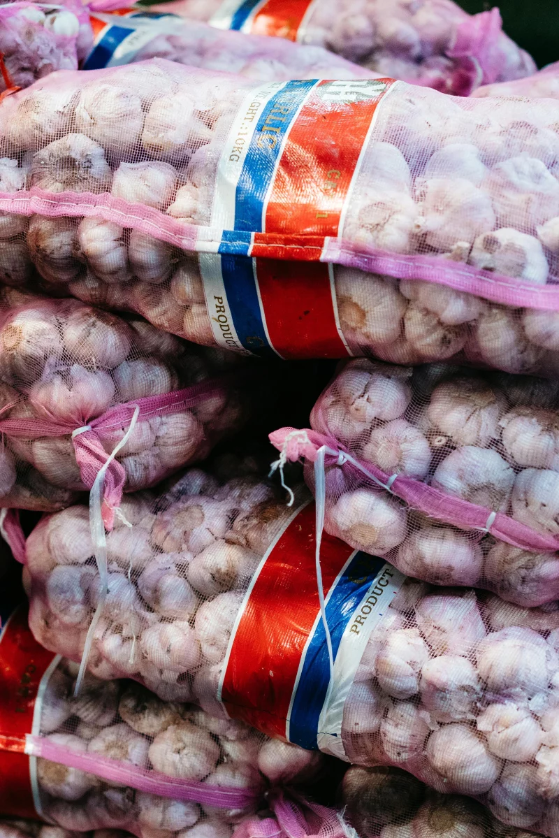 Stacks of bulk garlic contained in mesh bags, typical of market or wholesale settings.