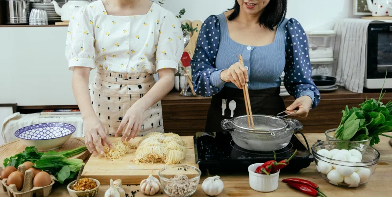 Two women cooking a noodle dish with fresh ingredients on a kitchen counter.