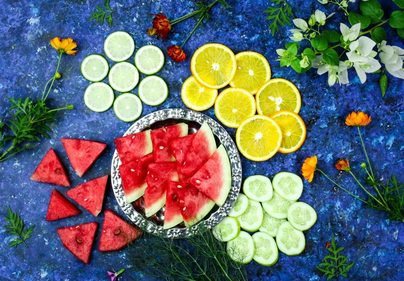 Colorful flatlay of sliced watermelon, orange, lime, and cucumber on a blue surface with flowers.