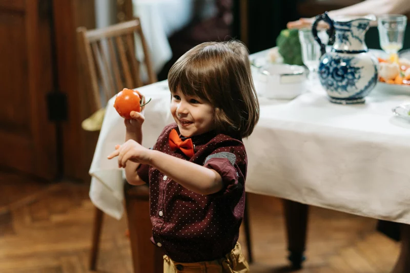 A cheerful child holds a tomato with a smile indoors, creating a warm, homely atmosphere.