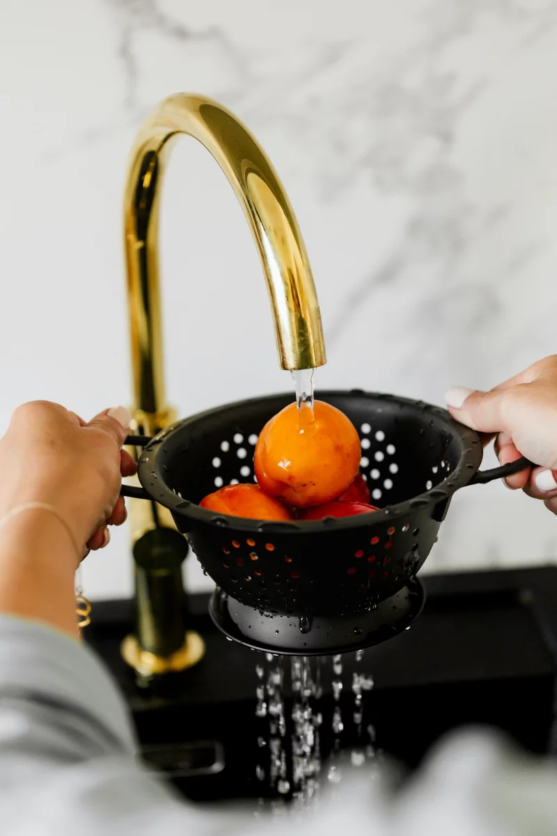 Close-up of hands washing oranges in a black colander under a gold faucet.