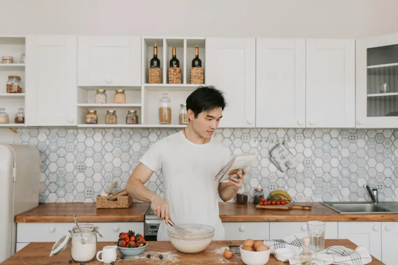 Young Asian man preparing a meal in a stylish kitchen, reading a recipe while mixing ingredients.