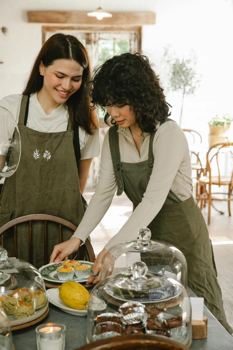 Two women in aprons arranging pastries on a café table, exuding warmth and hospitality.