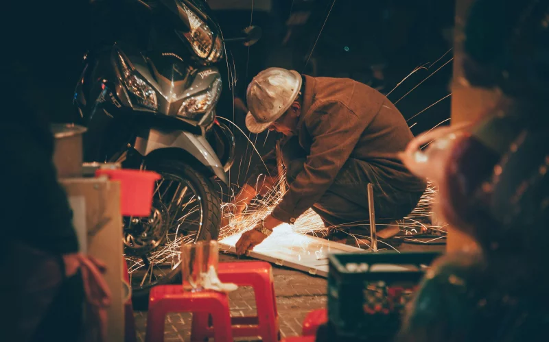 Artisan welding in a workshop, creating sparks beside a motorcycle, showcasing craftsmanship.