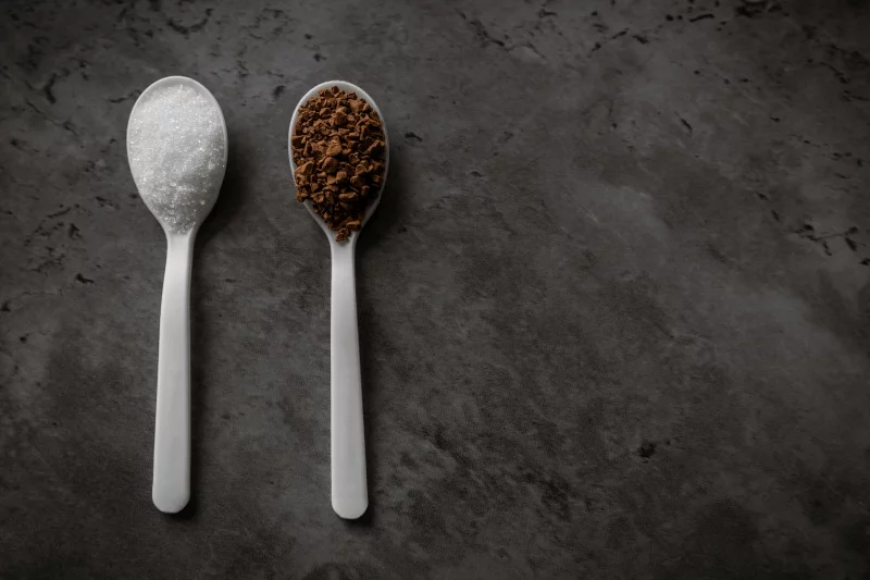 Close-up of sugar and coffee granules on spoons against a dark textured surface.