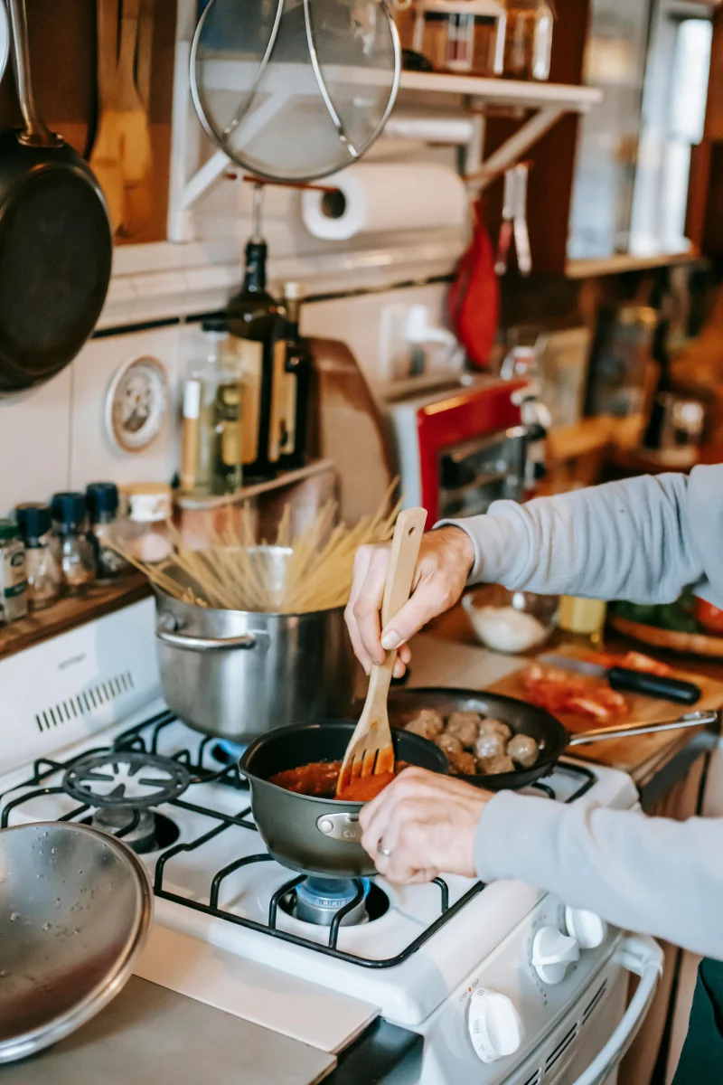 A person stirs homemade tomato sauce while cooking pasta and meatballs on a stove.