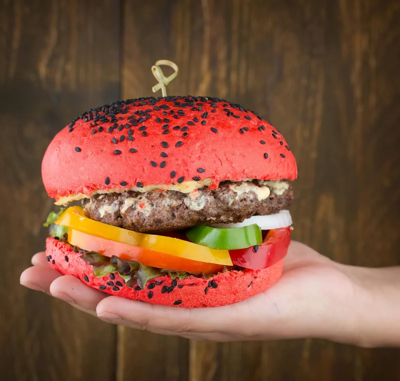 Colorful gourmet burger with red sesame bun and fresh vegetables held in hand.