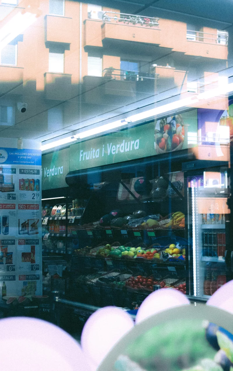 A vibrant produce aisle in a grocery store viewed through a reflective window, showcasing fresh fruits and vegetables.