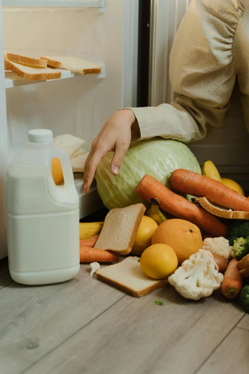 A person picks up fresh vegetables and bread from a refrigerator floor, highlighting kitchen mishap.