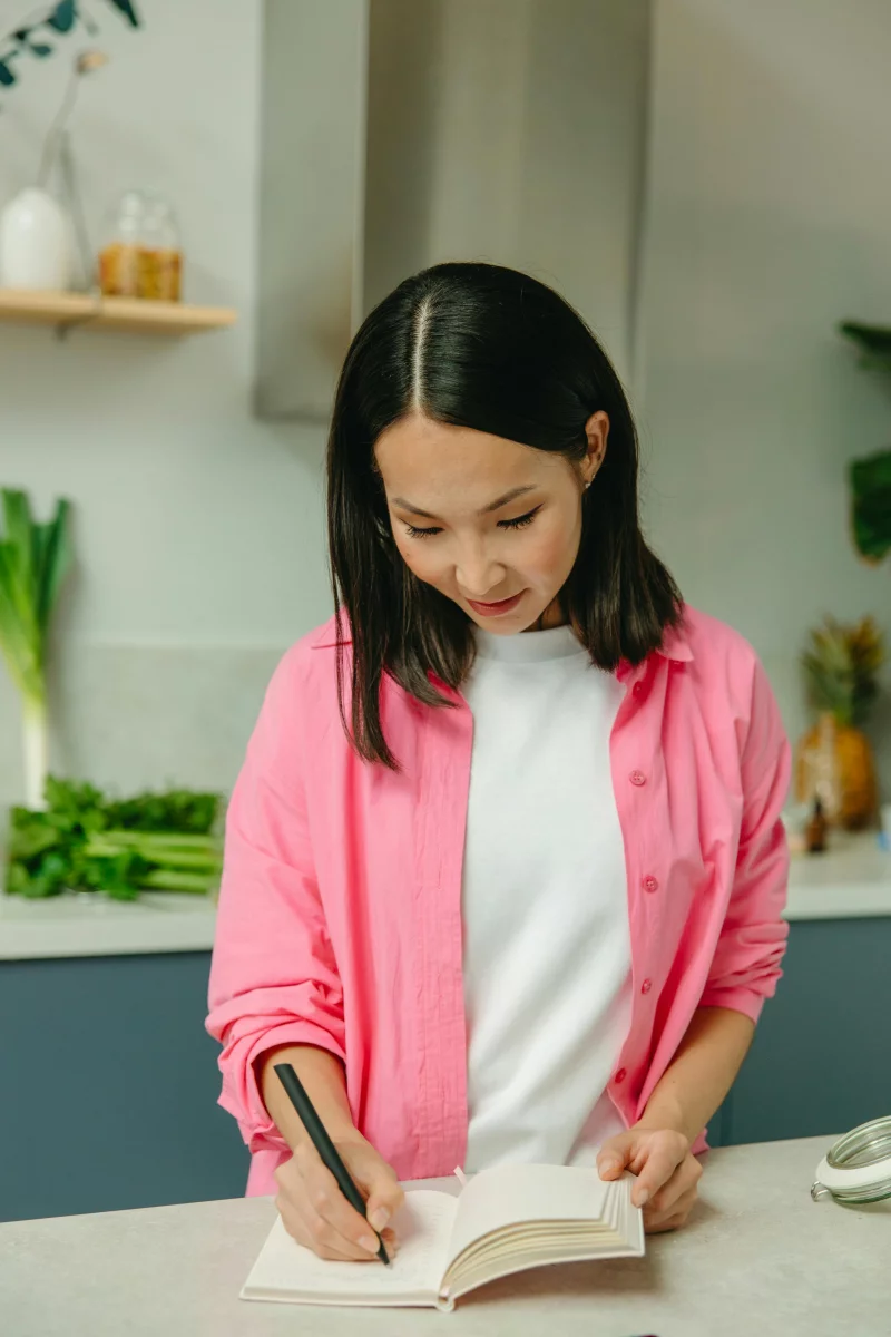 Asian woman in a pink overshirt writing in a notebook in a modern kitchen setting.