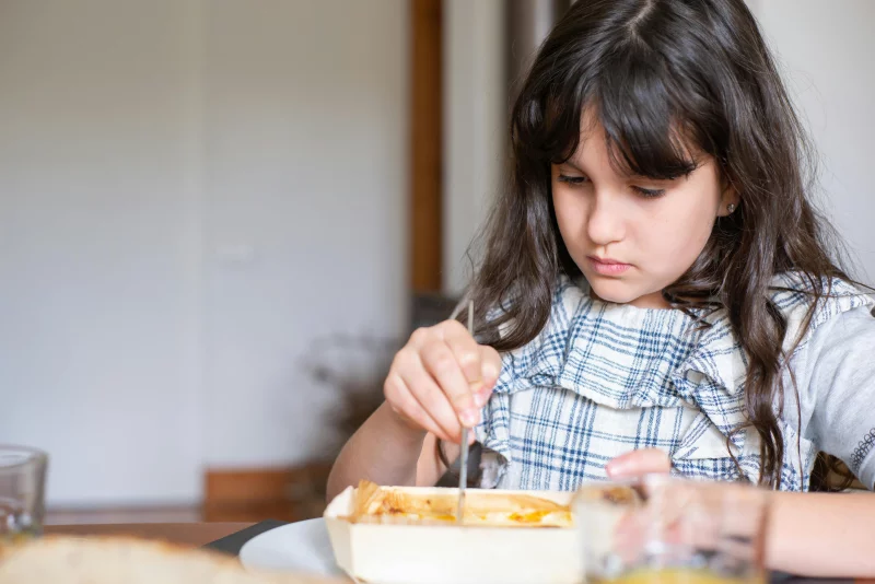 A young girl focuses on her meal at a dining table indoors.