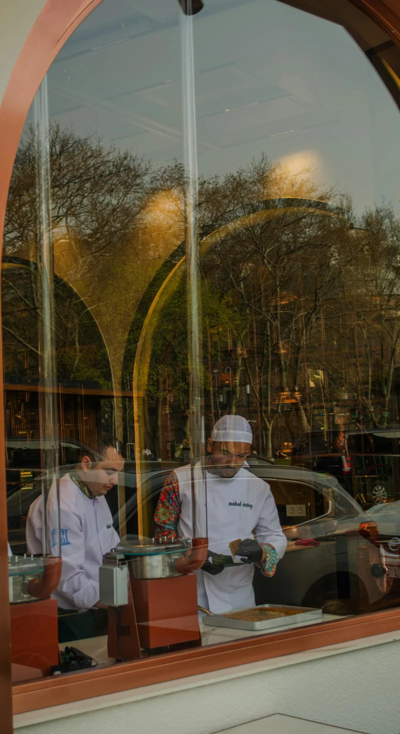 Chefs prepare meals in a bustling urban café with a reflective window view.
