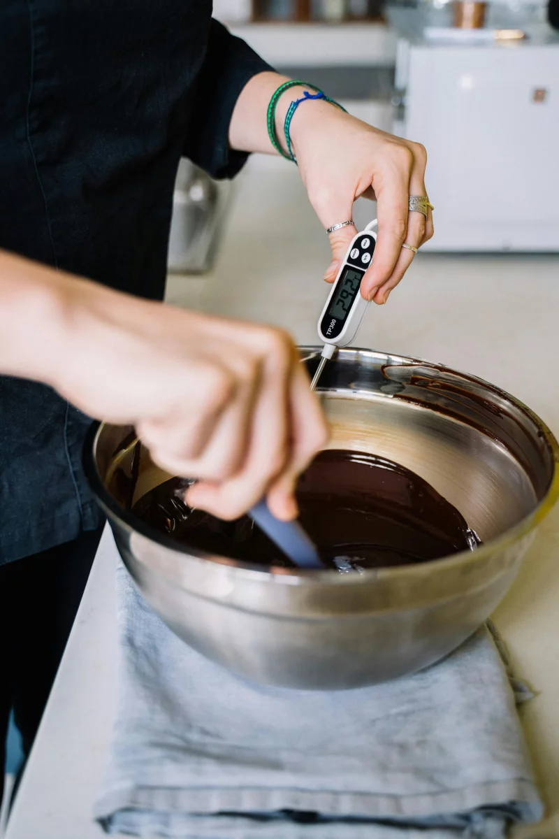 Close-up of hands tempering chocolate with digital thermometer in a kitchen.