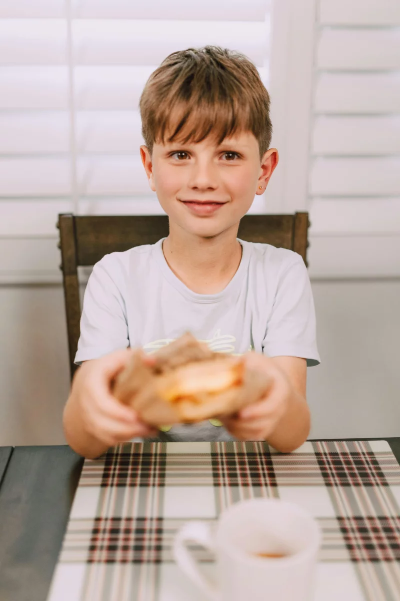 Cheerful boy holding a homemade cheeseburger at a dining table with a warm smile.