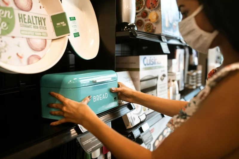 A woman shopping for a turquoise bread bin in a store while wearing a face mask.