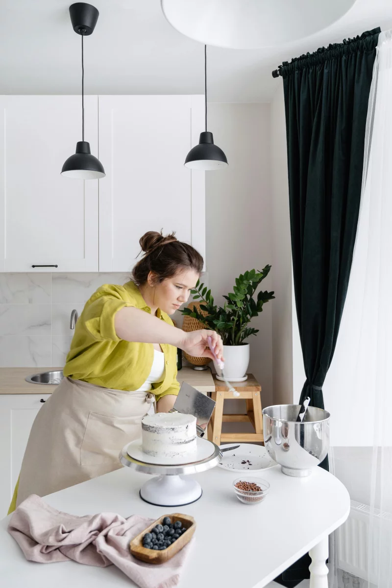 A woman in an apron decorates a cake in a bright, modern kitchen, emphasizing culinary art.