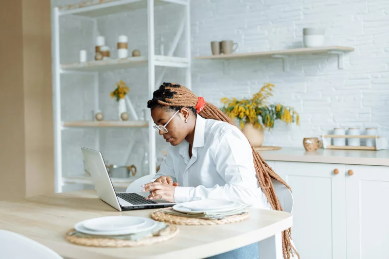 Young woman with braids working from home on a laptop in a bright, modern kitchen setting.