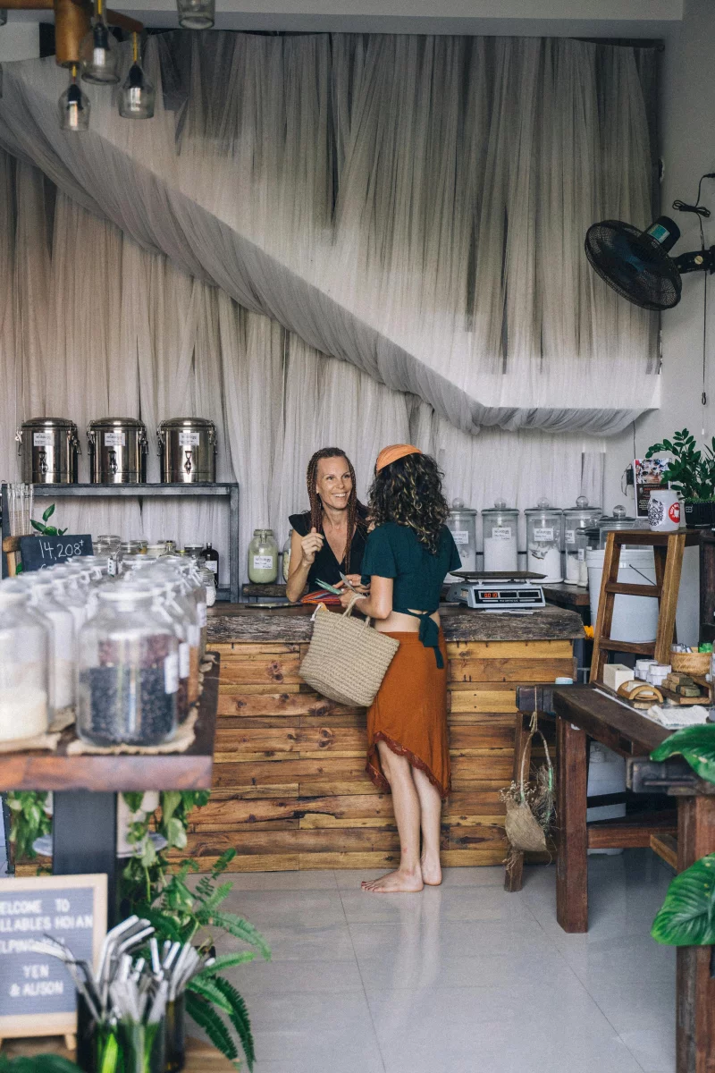 Two women interacting at a zero waste store counter, surrounded by refillable containers.