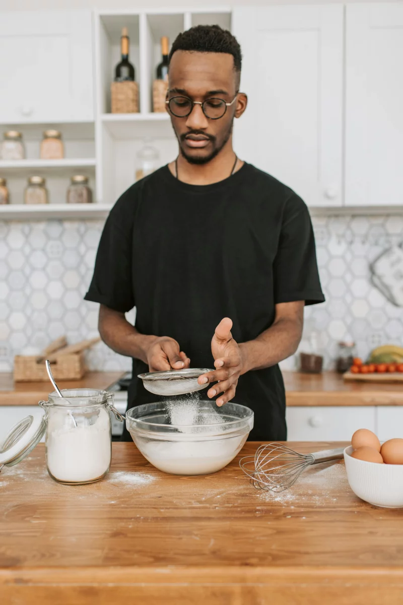 African American man sifts flour using a fine strainer in a stylish kitchen setting.