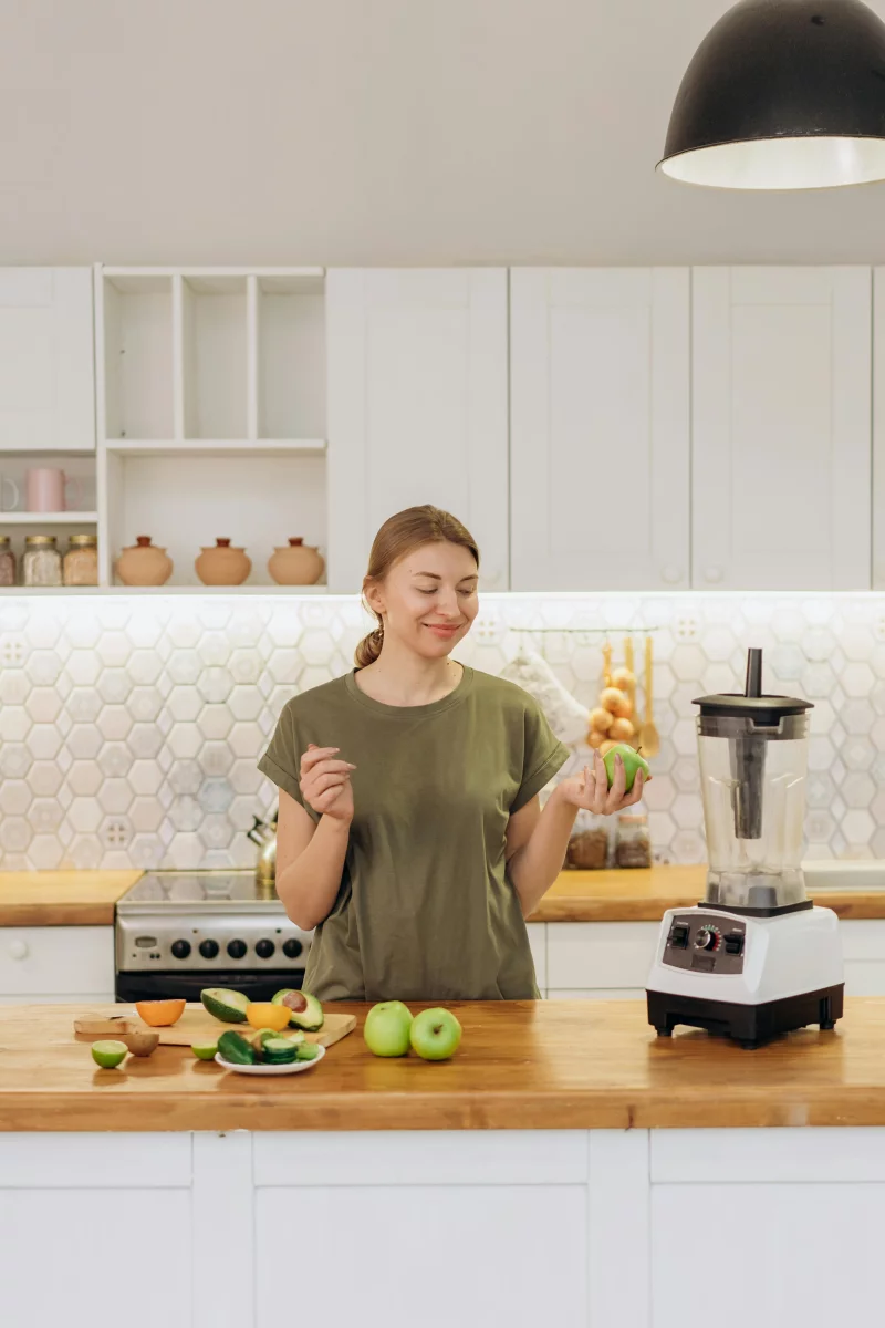 Smiling woman holding an apple in a stylish kitchen with blender and fresh fruit on the counter.