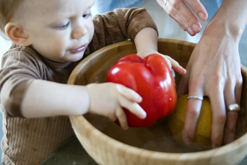 Child assisting an adult in washing vegetables in a wooden bowl, promoting healthy home activities.