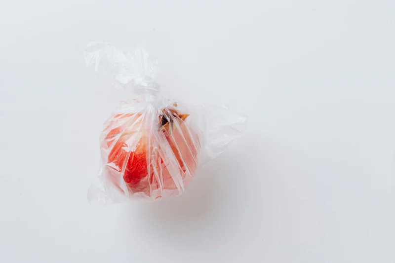 Minimalist photo of a fresh pomegranate wrapped in a clear plastic bag on a white background.