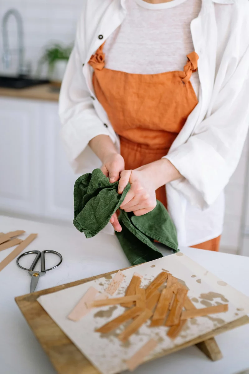 A woman wearing an apron dries wooden sticks with a green cloth in a kitchen.