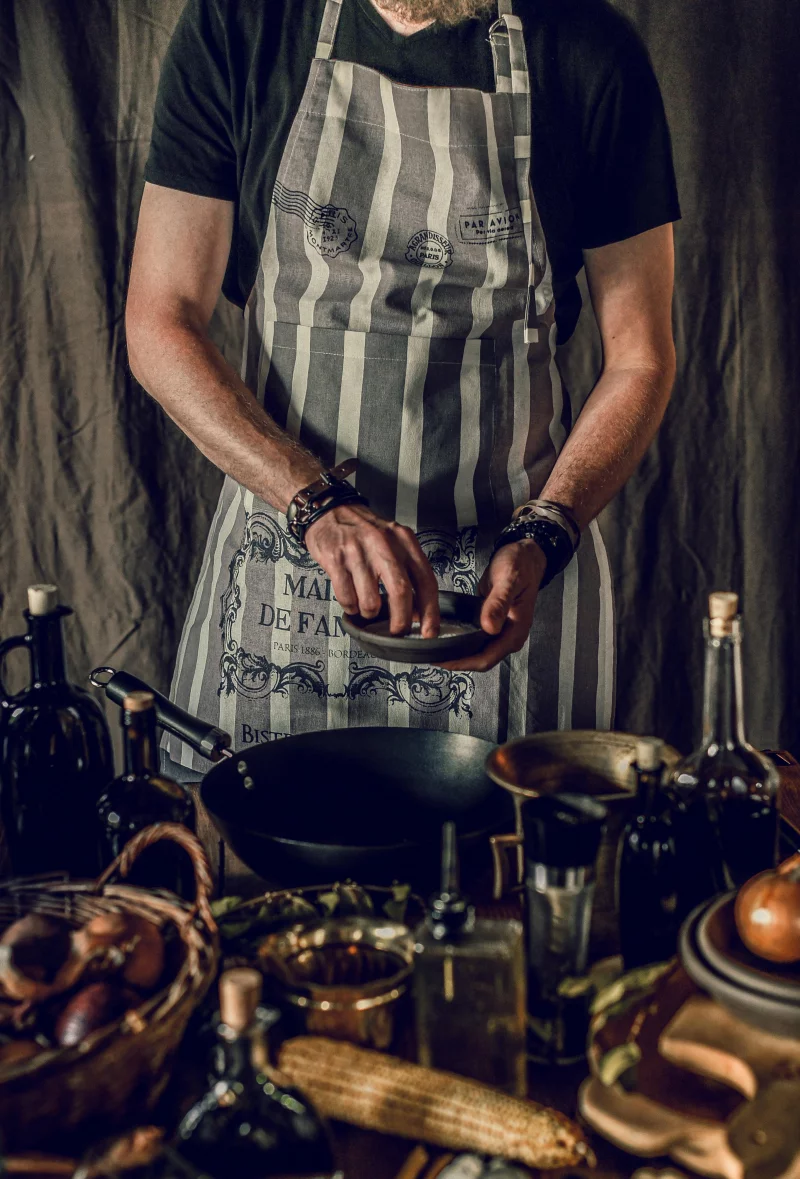 Chef seasoning a pan in a rustic kitchen with fresh ingredients.