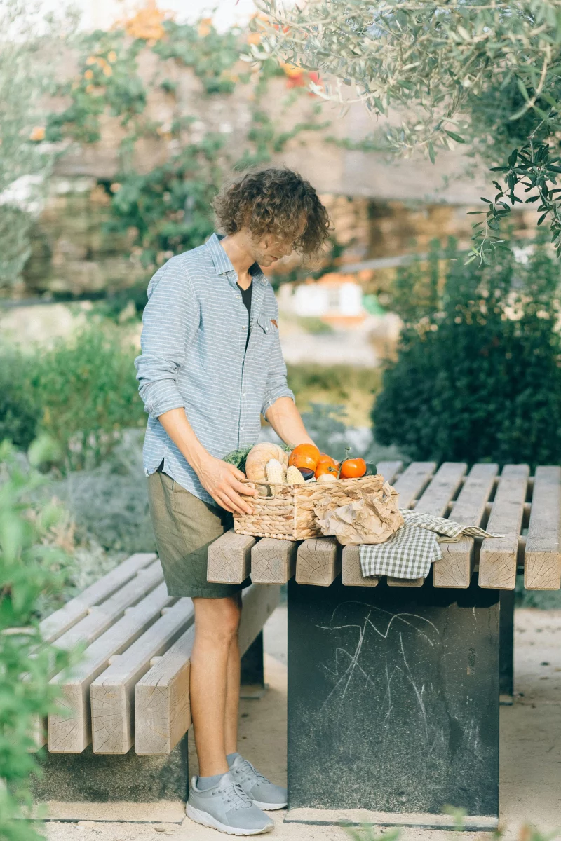 Person arranging fresh vegetables in a basket on a table in a garden setting.