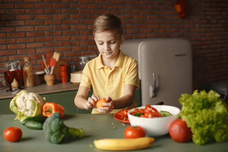 Confident kid peeling fresh orange sitting at table with assorted vegetables and fruits near salad bowl during vegetarian meal preparation in loft kitchen with brick walls