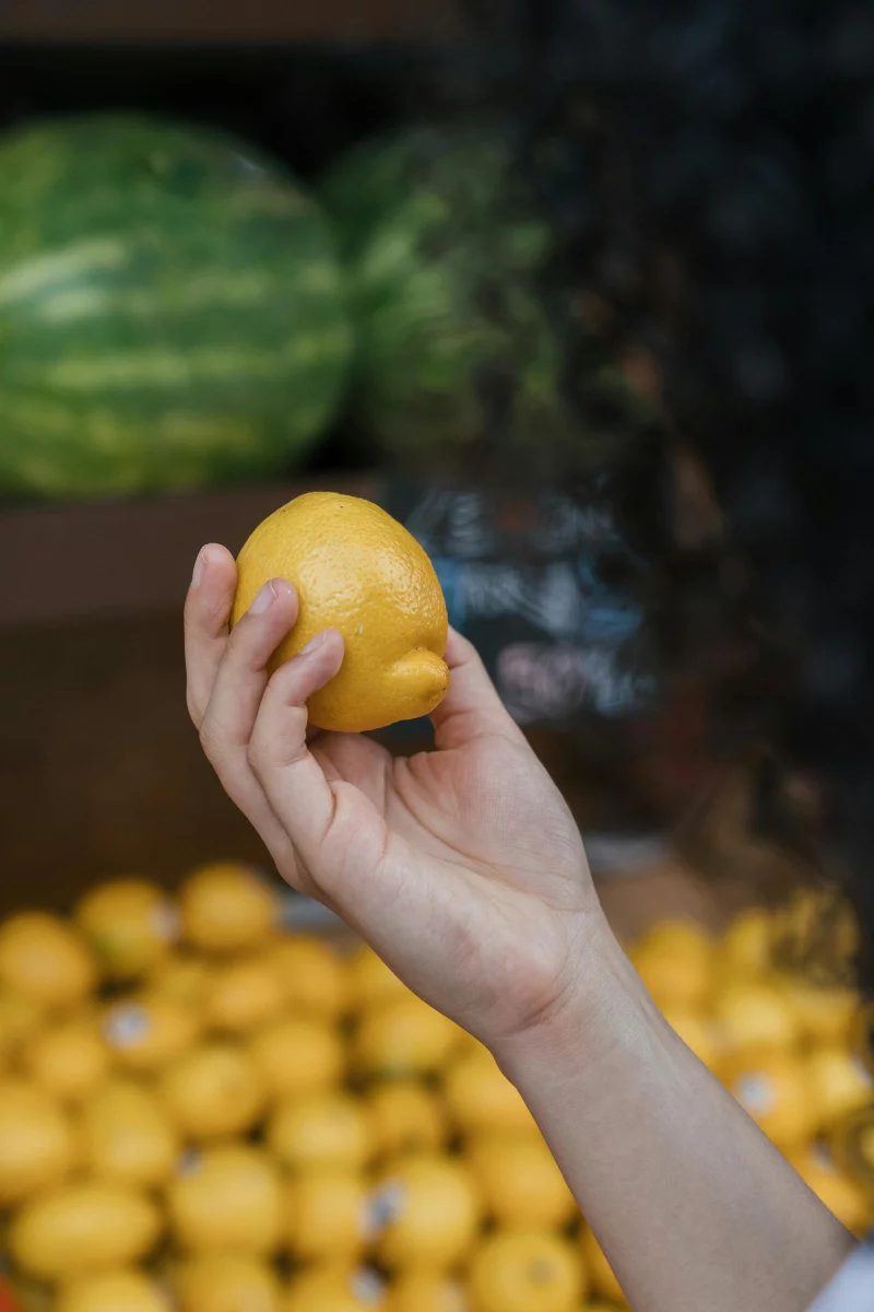Close-up of a hand holding a fresh lemon at a market with watermelons in the background.