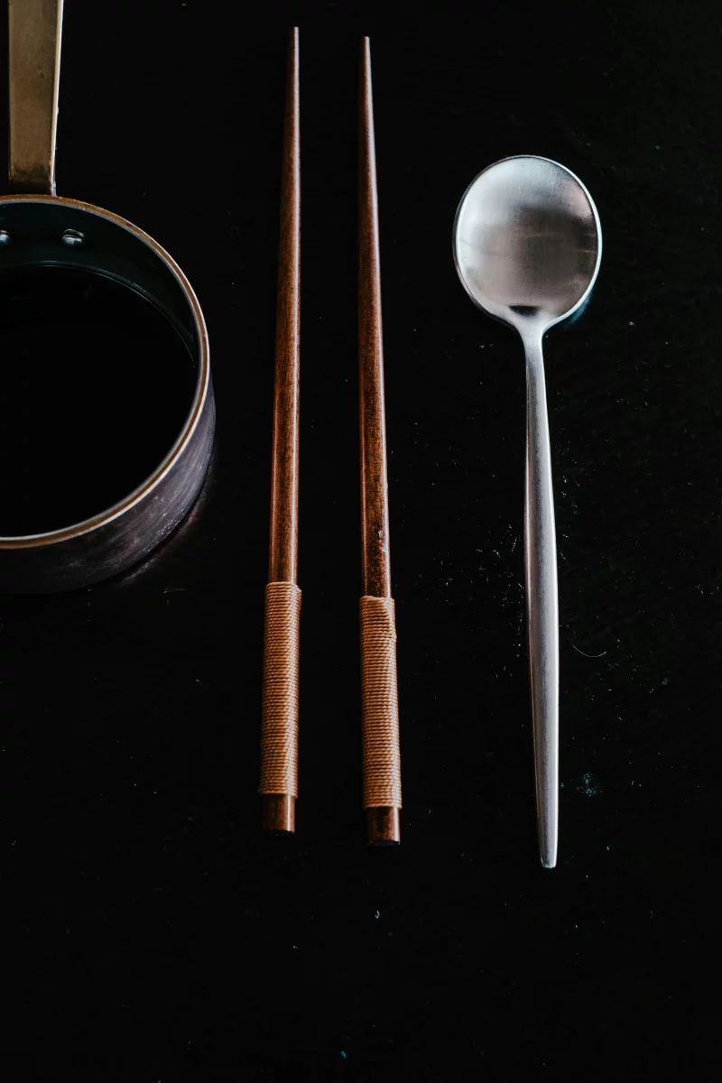 Minimalist display of kitchen utensils including chopsticks and spoon on a dark surface.