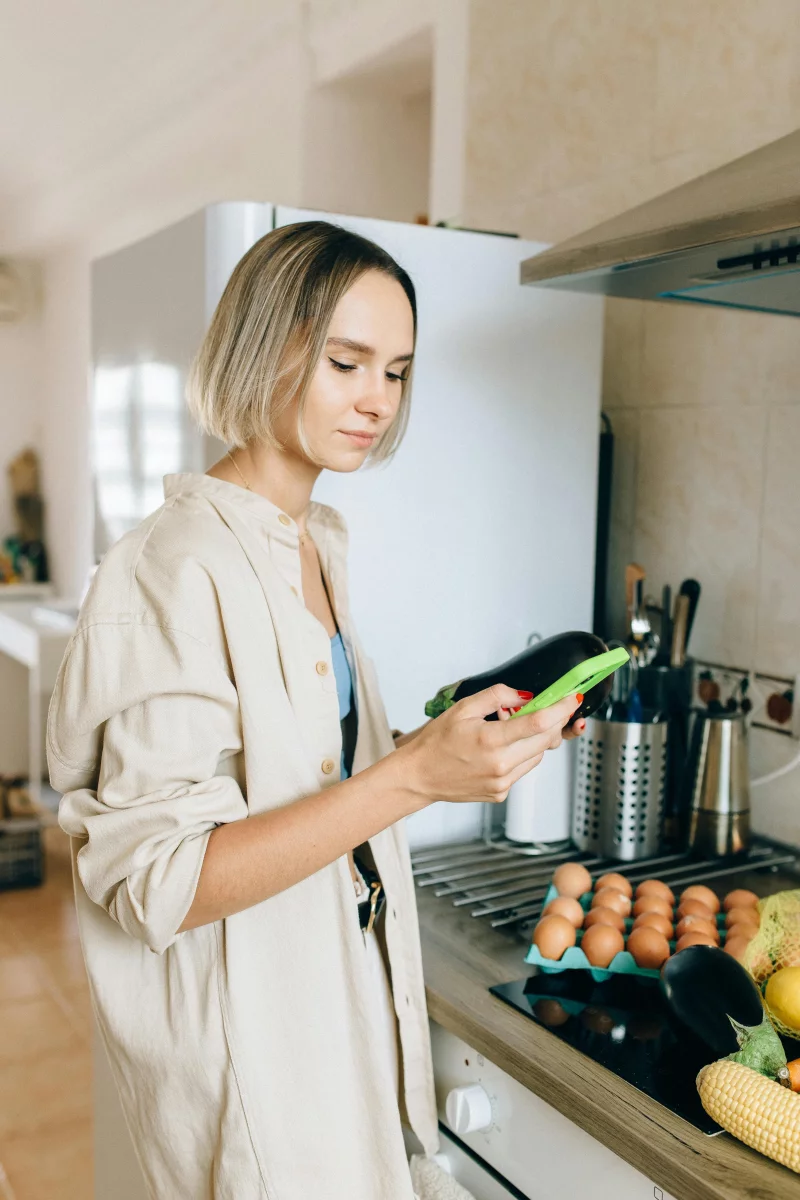 Woman browsing recipes on phone while cooking vegetables and eggs in a bright kitchen.