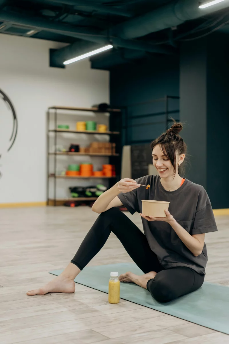A smiling woman sitting on a mat enjoying a healthy meal indoors after exercise.