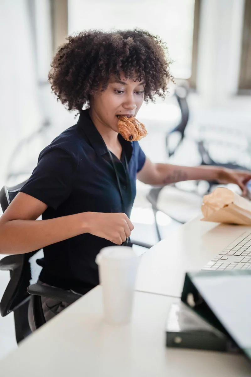 Young African American woman multitasking by eating a pastry while working at her office desk.