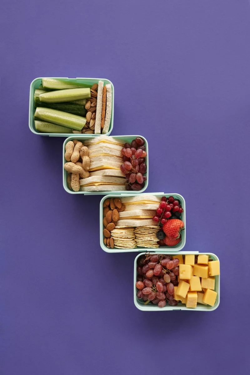 Top view of assorted healthy snacks in lunch boxes on a purple backdrop.